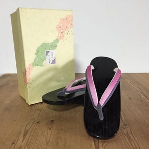 Könnte beinhalten: Ein Paar schwarze, lackierte Holz-Zori-Sandalen mit rosa und grauen Stoffriemen, begleitet von einer rechteckigen Aufbewahrungsbox mit hellgelbem Hintergrund, dekorativen grünen und pfirsichfarbenen abstrakten Formen und japanischem Text.