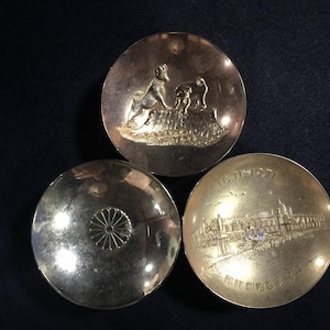 May include: Three antique silver-toned metal dishes. One dish features a relief of a dog and a smaller animal. Another has a floral design, and the third has a building and text. The dishes are circular.