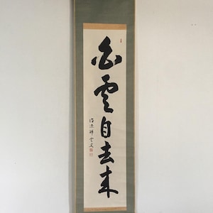 May include: A vertical wall hanging scroll featuring black ink calligraphy on a cream-coloured paper background. The scroll is framed by a sage green fabric border with light tan accents at the top and bottom. Red seal stamps are visible.