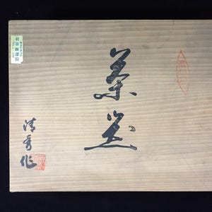 May include: A rectangular light brown wooden box featuring prominent black Japanese calligraphy characters centered on the lid. A small rectangular label with Japanese text and a red stamp are visible on the left side, with a red seal on the right.