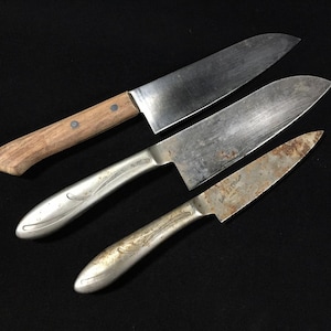 Może przedstawiać: Trzy zabytkowe noże kuchenne z metalowymi ostrzami. Jeden nóż ma drewnianą rękojeść, a dwa pozostałe srebrne. Ostrza noszą ślady zużycia i wieku, z widoczną rdzą. Noże ułożone są na czarnej powierzchni.