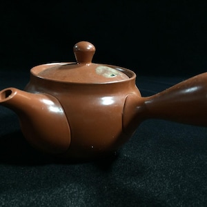 May include: A brown ceramic teapot with a side handle and a lid. The teapot has a spout and a rounded body. The handle is a large, open loop. The teapot is set against a dark background.