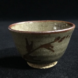 May include: A small, round ceramic bowl with a speckled, muted gray-green glaze and a dark brown rim. The exterior features a hand-painted, abstract dark brown branch design. The base of the bowl is unglazed, revealing a rough, light beige texture.