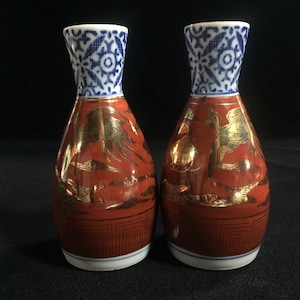 May include: Two Japanese sake bottles. The bottles are red with gold accents and a geometric pattern at the base. The necks are white with a blue and white floral design. The bottles are on a black surface.