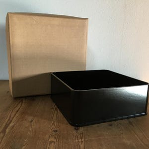 Peut inclure: Un récipient carré noir à dessus ouvert avec des coins arrondis est placé devant une grande boîte carrée de couleur beige. Le récipient noir a une finition lisse et réfléchissante, tandis que la boîte beige a une texture mate.