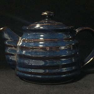 May include: A ceramic teapot featuring a deep blue glaze with horizontal ribbed textures and dark brown accents along the ridges. The teapot includes a matching lid with a small knob handle, a curved spout, and a sturdy side handle for pouring.