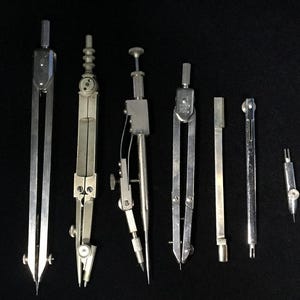 Könnte beinhalten: Eine Sammlung antiker silberner Zeichenzirkel und -teiler, angeordnet auf einem schwarzen Hintergrund. Die Werkzeuge variieren in Größe und Design und verfügen über scharfe Spitzen und verstellbare Arme, die für präzises Zeichnen und Messen in der Geometrie und im technischen Zeichnen verwendet werden.