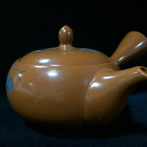 May include: A brown ceramic teapot with a side handle and a matching lid. The teapot features a smooth, rounded body and a short, tapered spout. A small, delicate blue leaf design is painted on the side of the pot, adding a subtle detail to the solid brown surface.