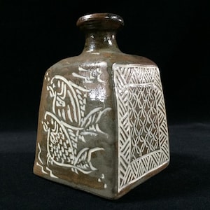 Peut inclure: Une bouteille en céramique marron avec un corps carré et un col étroit. La bouteille présente des motifs de poissons blancs et géométriques. Le vernis a une finition brillante, et le fond est noir.