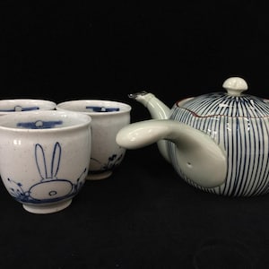May include: A ceramic teapot with a side handle and a blue striped pattern, accompanied by two matching ceramic cups featuring a blue hand-painted rabbit design on a speckled white background. The teapot has a lid with a small knob handle.