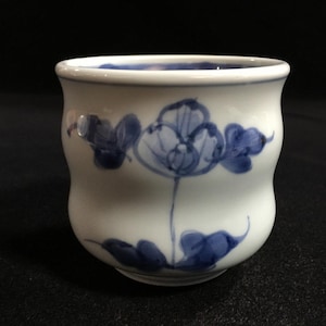 May include: A white ceramic mug with a black handle and the text "But First, Coffee" in a bold, black font. The mug is set against a blurred background, suggesting a cozy setting.