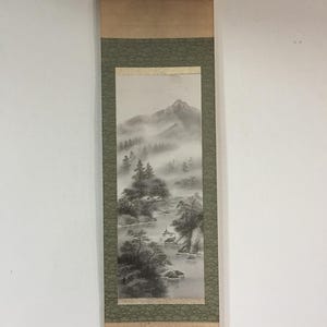 Könnte beinhalten: Eine vertikale Hängerolle mit einer monochromen Tuschezeichnung einer nebligen Berglandschaft mit Kiefern, einem Fluss und kleinen Gebäuden. Das Kunstwerk ist auf einem grünen, gemusterten Stoffrand mit beigen Enden montiert.