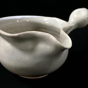 May include: A light gray ceramic creamer or pouring vessel with a glossy crackle glaze finish. The piece features a rounded body, a wide spout for pouring, and a single bulbous handle extending from the side, showcasing a fine network of lines.