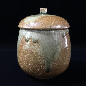Op de afbeelding: Een keramische pot met deksel, voorzien van een gespikkeld bruin en beige glazuur met een lichtgroen, craquelé glazuur dat vanaf de bovenkant naar beneden druipt. De pot heeft een ronde, bolvormige vorm en een kleine knop op het deksel.