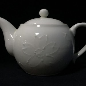 May include: A white ceramic teapot with a floral design embossed on the body. The teapot has a curved spout, a rounded handle, and a lid with a small knob. The teapot is set against a dark background.