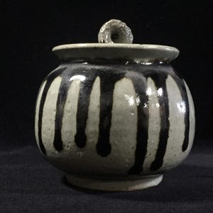 Può includere: Un barattolo in ceramica rotondo con coperchio, caratterizzato da una smaltatura di base grigio chiaro e motivi a goccia verticali marrone scuro. Il barattolo presenta un'ampia apertura, un collo corto e un piccolo pomello testurizzato sul coperchio.
