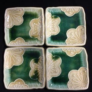 May include: Four square ceramic plates with a green and cream floral design. The plates have a dark green centre with a cream-coloured floral pattern in each corner. The plates are arranged in a square formation.