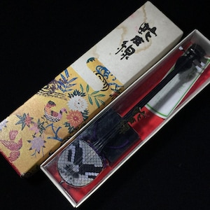May include: A miniature Japanese shamisen, a three-stringed lute, in a decorative box. The box has a gold background with floral and bird designs. The instrument is black with a patterned soundboard.