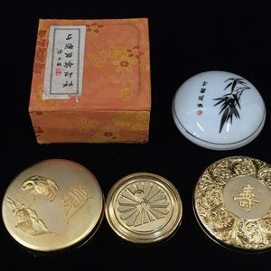 Pode incluir: Uma coleção de quatro recipientes redondos, incluindo uma caixa laranja com padrão floral, uma tampa de cerâmica branca com ilustrações de bambu preto e três estojos de metal dourado com desenhos em relevo de garças, flores e caracteres.