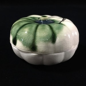 以下が含まれることがあります： カボチャやヒョウタンの形をした蓋付きの丸い陶器製容器。蓋は白いベースに緑色の釉薬が塗られ、細かいひび割れ模様があり、上部には小さな青い取っ手が付いています。下部は無地の白で、リブ状の質感があります。