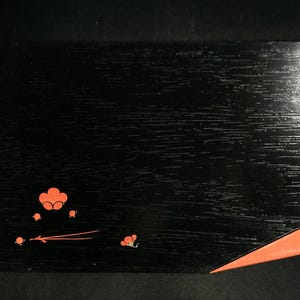 以下が含まれることがあります： 黒い長方形の漆塗りの箱で、表面はテクスチャ加工されています。箱の左下隅にはオレンジ色の様式化された花の模様があり、右下隅にはオレンジ色の三角形があります。箱は、おそらく保管または表示に使用されます。