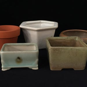 May include: A collection of five ceramic and terracotta plant pots in various shapes and colors. The set includes a round terracotta pot, a white hexagonal pot, a rectangular light blue pot, a square beige pot, and a round brown pot with studs.