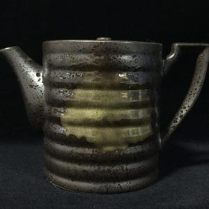 May include: A cylindrical ceramic teapot featuring a dark, speckled metallic glaze with horizontal ridges along the body. A central band of muted yellow glaze contrasts with the dark finish. The teapot includes a curved spout and a handle.