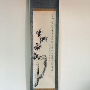 May include: A vertical scroll painting featuring a bamboo and rock ink wash illustration. The artwork is primarily black and white, with a focus on the detailed depiction of bamboo stalks and leaves. The scroll has a dark blue border and includes Chinese calligraphy.