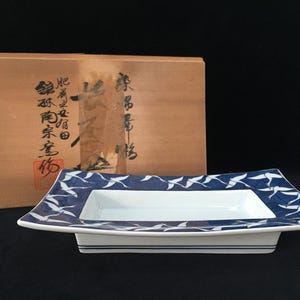以下が含まれることがあります： 白い中央部と、飛んでいる白い鶴の繰り返し模様が描かれた幅の広い青い縁を持つ、長方形の陶磁器製盛り皿。この皿には、黒い日本語の書が書かれた明るい茶色の木製収納箱が付属しています。