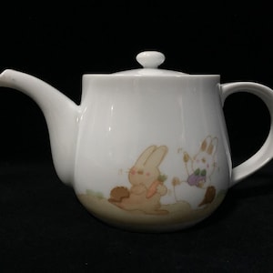 Op de afbeelding: Een witte keramische theepot met een rond lichaam, een gebogen tuit en een bijpassend deksel met een kleine knop. De onderkant van de theepot is versierd met een illustratie in zachte kleuren van twee konijntjes, een lichtbruine en een witte.