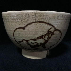 May include: A beige ceramic bowl with a cracked glaze effect. A brown illustration of an animal is inside a brown cloud-shaped design. The bowl has a small, off-white pedestal base. The bowl is likely for serving food.