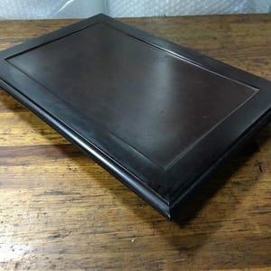 May include: A rectangular wooden tray with a dark brown finish and a raised border frame. The surface is smooth and flat, featuring a deep espresso color with subtle wood grain visible across the central panel and the beveled edges.