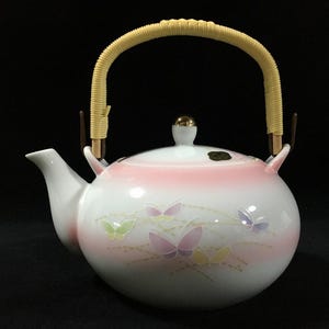 May include: A white ceramic teapot with a pink band and butterfly design. The teapot has a woven handle and a gold-colored knob on the lid. The spout is white and curved.