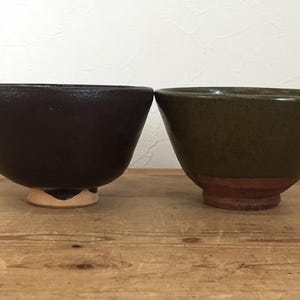 May include: Two ceramic bowls with a conical shape. The bowl on the left has a dark brown, textured matte finish and a light-colored base. The bowl on the right features a smooth olive green glaze on the upper portion and an exposed reddish-brown clay base.