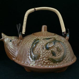 Op de afbeelding: Een bruine keramische theepot in de vorm van een schildpad met een textuur op het schild, een puntige tuit en een deksel. De theepot heeft een gebogen handvat omwikkeld met lichtgekleurd rieten materiaal en donkerbruine decoratieve accenten.