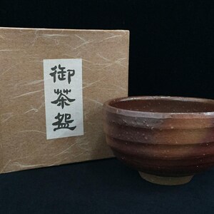 Può includere: Una ciotola da tè in ceramica marrone con una consistenza esterna a coste e una finitura maculata, accompagnata da una scatola quadrata color marrone chiaro con un'etichetta bianca con caratteri calligrafici giapponesi neri 御茶盤.