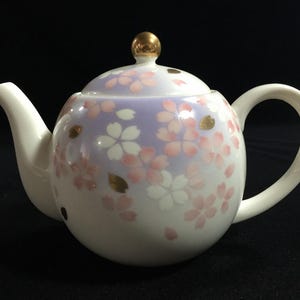 May include: White ceramic teapot with a rounded body, a curved handle, and a spout. The teapot is decorated with pink cherry blossom designs and gold accents on a lavender and white background. The lid is topped with a gold knob.