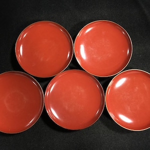 May include: Five round, red plates with a glossy finish and a thin gold rim. The plates are arranged on a dark surface, showcasing their simple, elegant design. These plates are suitable for serving appetizers or desserts.