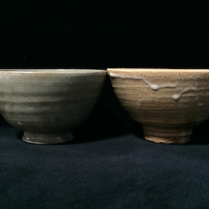 May include: Two ceramic bowls with a rustic aesthetic. The bowl on the left is a muted gray-green, while the one on the right is tan with white drips. Both have a footed base and textured, ridged sides.