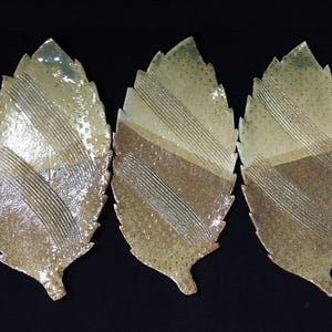 May include: Three ceramic leaf-shaped plates. The plates have a textured surface with a color gradient from light beige to brown. Diagonal lines are etched across the surface. The plates are arranged on a black background.