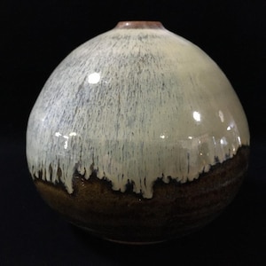 May include: A spherical ceramic vase featuring a glossy off-white glaze that drips down over a dark brown base. The upper portion of the vase has a smooth, rounded shape with a small circular opening at the top, while the lower section displays a textured, earthy brown finish where the white glaze has settled in vertical streaks.