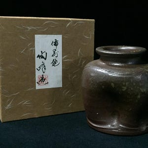 以下が含まれることがあります： 広い口と質感のある不均一な表面を持つダークブラウンの陶器の壺。その横には、黒い日本語の書道と赤い四角い印が押された白い縦長のラベルが貼られた、正方形のベージュ色の段ボール箱が置かれています。
