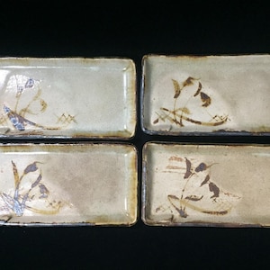 May include: A set of four rectangular ceramic plates with a light beige, speckled glaze. Each plate features a hand-painted brown botanical motif of leaves and stems in one corner, with dark, uneven edges and a slightly textured, rustic finish.