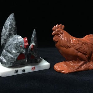 以下が含まれることがあります： 黒い中国語の文字と赤い印章が施された白い長方形の台座に乗った、灰色と白の斑点のある石の鳥の彫刻と、羽のディテールが細かく施された赤茶色の粘土製の雄鶏の置物。