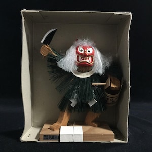 May include: A Japanese wooden figure of a demon with a red face, white hair, and a green outfit, holding a knife and a bucket. The figure is inside a cardboard box.