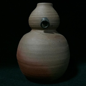 May include: A gourd-shaped ceramic vessel with a matte, earthy brown finish and a subtle reddish-orange gradient near the base. The vessel features a narrow neck, a rounded body, and a small, dark, circular spout protruding from the upper section.