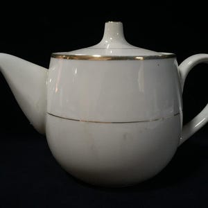 Op de afbeelding: Een witte keramische theepot met een rond lichaam, een gebogen handvat en een puntige tuit. Het deksel heeft een kleine knop aan de bovenkant. Een dunne, metallic gouden band loopt rond de rand van het deksel en het midden van de theepot.