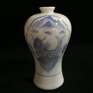 Op de afbeelding: Een witte porseleinen vaas met een smalle hals en een ronde buik. Het oppervlak is voorzien van een blauw geschilderd ontwerp van een koi-karper die in water zwemt onder gestileerde bergen en wolken, omlijst door een decoratieve rand.
