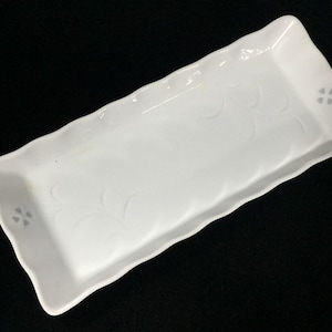 May include: A rectangular white ceramic serving tray with a wavy, scalloped edge. The surface features a subtle embossed leaf pattern and small four-petal flower motifs at each end of the tray, creating a clean and simple aesthetic.