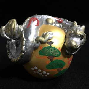 Könnte beinhalten: Dekorative, handbemalte japanische Keramik-Drachenfigur. Der Drache ist silber- und goldfarben und umschlingt eine goldfarbene Kugel mit bemalten grünen Bäumen und weißen floralen Akzenten. Die Figur steht vor einem dunklen Hintergrund.
